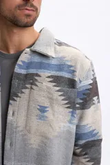 Sobrecamisa de hombre con estampado tribal en tonos gris claro, azul y marrón, de manga larga, cuello clásico, bolsillos frontales con solapa y cierre de botones.