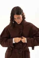 Trench color chocolate con cuello alto, cierre frontal con botones y cinturón ajustable en la cintura. Presenta mangas largas con puños ajustables mediante tiras y hebillas, y bolsillos frontales.