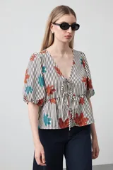 Blusa de silueta relajada con estampado de rayas verticales y motivos florales en tonos naranja y turquesa. Presenta escote en V con lazos frontales regulables, mangas cortas con leve volumen y detalle de frunce bajo el busto que aporta movimiento.