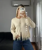 Cárdigan de tejido de punto color crema, con cuello redondo, mangas largas y cierre frontal con botones metálicos. Presenta detalles de flecos en los puños y en el ruedo.
