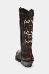 Bota estilo cowboy de cuero marrón oscuro con bordados florales en tonos claros y rojizos. Presenta tacón ancho en bloque, puntera afilada y tiradores laterales para facilitar el calce.