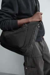 Bolso de hombro tipo hobo o media luna, confeccionado en tejido técnico negro. Presenta un bolsillo frontal con cierre y una correa ajustable para llevar al hombro.