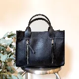 Cartera tipo tote de cuero negro, con estructura firme, doble asa superior y dos bolsillos frontales verticales con cierre metálico. Incluye correa desmontable.