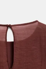 Blusa marrón de cuello redondo y manga larga, con volantes horizontales superpuestos y bajo asimétrico. Cuenta con detalle de abertura y botón en la espalda.