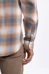 Camisa de manga larga con estampado de cuadros en tonos gris, beige y blanco. Presenta cuello abotonado, cierre frontal con botones y un bolsillo en el pecho.
