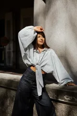 Camisa de algodón con diseño a rayas verticales en tonos blanco y gris. Presenta un corte oversize con mangas abullonadas y un nudo frontal que permite ajustar la prenda a la cintura.