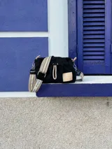Cartera bandolera de nylon color negro con detalles en charol. Presenta un diseño fruncido con cordones decorativos combinados, grifa de marca en material sintético y correa regulable e intercambiable con logo estampado.