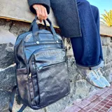 Mochila urbana de cuero sintético negro, con múltiples compartimentos frontales con cierre, bolsillos laterales y compartimento interno para notebook. Cuenta con correas ajustables y diseño funcional para uso diario.