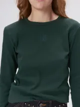 Remera de manga larga con cuello redondo, confeccionada en tejido acanalado de color verde oscuro. Presenta un logo bordado en el centro del pecho y costuras a contratono.