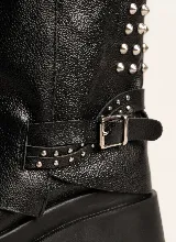 Bota corta de cuero negro con diseño de polaina superpuesta, detalle de tachas metálicas en el lateral y correa con hebilla ajustable. Cuenta con suela track de goma y taco cuadrado.