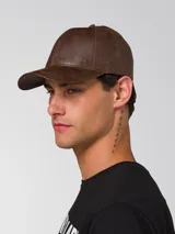 Gorro tipo gorra de béisbol con visera curva, confeccionado en cuero sintético con acabado texturizado. Presenta un pequeño aplique metálico con el logo de la marca en el lateral frontal y cierre metálico ajustable en la parte posterior.