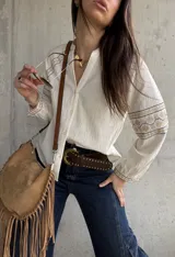 Camisa de inspiración boho de color crudo, con mangas largas abullonadas y puños decorados con detalles bordados geométricos en tonos tostados y dorados. Presenta cuello en V con cierre frontal de botones.