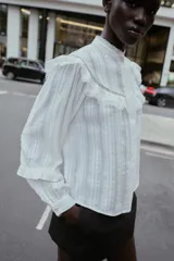 Blusa blanca de manga larga con cuello redondo, confeccionada con detalles de bordados y volantes a tono. Presenta cierre frontal con botones ocultos y puños ajustados.