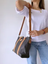 Cartera de cuero negro con detalles en cuero color suela, incluyendo asas fijas y correa larga ajustable. Presenta una etiqueta frontal con logo grabado y cierre superior.