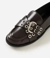 Mocasín femenino de cuero sintético color marrón oscuro, con punta redondeada y suela plana. Presenta un diseño con tiras cruzadas en el empeine decoradas con ojales metálicos y una hebilla ajustable.