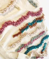 Sweater tejido a mano en punto jersey de 100% lana merino, color blanco con detalles texturizados en punto bucle de tonos rosados, mostaza, marrón y gris, formando líneas onduladas en el frente y las mangas. Cuenta con cuello redondo base y puños largos acANALados.