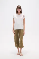 Remera blanca de algodón con mangas cortas y cuello redondo. Presenta un bordado floral en tonos rosados y rojizos en la zona del hombro derecho.
