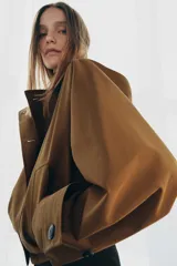 Trench corto color verde militar, con cuello solapa y capucha. Tiene manga larga con trabillas y botón en los puños, bolsillos delanteros y bajo asimétrico. Cierre frontal con botones. El tejido es repelente al agua.