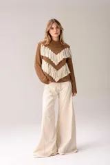 Sweater de punto color crema con cuello medio alto y silueta relajada. Presenta un diseño frontal con líneas en V decoradas con flecos largos de color marrón aplicados a mano.