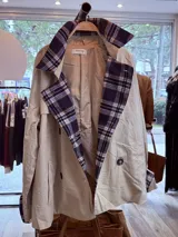 Gabardina de corte clásico en color verde militar, con solapas amplias y puños con detalles en estampado de cuadros escoceses en tonos azul, blanco y negro. Presenta cierre frontal cruzado con botones y bolsillos laterales.