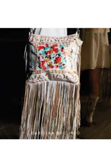 Cartera bandolera de cuero color blanco, decorada con flecos largos en tonos blancos y tostados, y una intervención artística pintada a mano con diseño abstracto en tonos rojos, celestes y dorados.