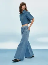 Camisa corta de denim azul claro, con cuello camisero, manga corta ancha y cierre frontal de botones blancos. Presenta un bolsillo de parche en el pecho izquierdo.