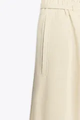 Pantalón ancho de tiro alto color beige claro, con cintura elástica, cordón ajustable y pinzas delanteras. Presenta bolsillos frontales y bolsillos de plastrón en la espalda. Incluye un cinturón del mismo tejido con apliques metálicos.