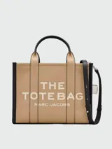 Cartera tipo tote bag Marc Jacobs modelo The Medium Leather Tote Bag, color marrón tostado con detalles en negro, confeccionada en cuero con las inscripciones "THE TOTE BAG" y "MARC JACOBS" estampadas en el frente. Cuenta con asas superiores y correa ajustable y desmontable para el hombro.