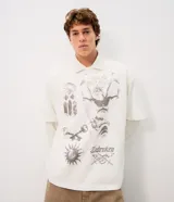 Remera oversize de algodón color blanco, con cuello polo y estampado frontal en tonos grises con gráficos de estilo gótico y vintage.