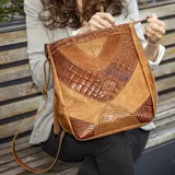 Bolso tipo morral de cuero vacuno color tostado y marrón, con correa larga ajustable y extraíble, y asa corta. Tiene dos bolsillos verticales en los laterales, forro interior de tela con bolsillo con cierre y bolsillo para el móvil.