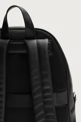 Mochila de diseño minimalista en color negro con acabado texturizado. Cuenta con un compartimento principal con cierre de cremallera, un bolsillo frontal con solapa y compartimento interior para dispositivos electrónicos. Incluye espalda acolchada, banda de sujeción para equipaje, asa de mano y correas de hombro ajustables.