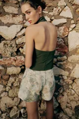 Chaleco verde militar con cuello halter, espalda descubierta y cierre frontal con cremallera oculta. Presenta detalles de falsos botones dorados y ribetes en contraste, con trabillas en los laterales.