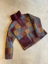 Buzo de cuero genuino con diseño de patchwork en tonos marrón, gris y bordó. Presenta cuello alto con cierre frontal parcial, bolsillo frontal tipo canguro y detalles de piel sintética en cuello, cierre y bolsillo.