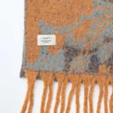 Bufanda de tejido suave con diseño jaspeado en tonos naranja y azul, terminada con flecos en los extremos y etiqueta de marca cosida.