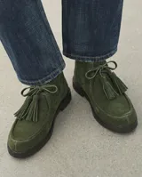 Botas cortas de gamuza color verde militar, con cierre de cordones de cuero y borlas colgantes en el empeine. Presentan punta redondeada y suela plana oscura.