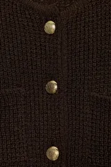 Chaqueta de punto texturizado color marrón oscuro, de corte cropped y cuello redondo. Presenta cierre frontal con botones dorados y dos bolsillos de plastrón decorativos con botón dorado en el pecho.