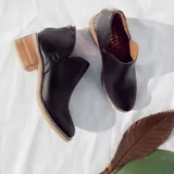 Botín de cuero vacuno color negro, con diseño de corte lateral, cierre metálico en el talón y taco foliado de madera en tono natural.