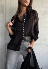 Camisa de color negro con cuello clásico y cierre frontal de botones blancos. Presenta mangas largas con detalles bordados en los puños.