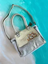 Cartera de cuero blanca y dorada con solapa texturizada tipo cocodrilo y detalles pintados a mano. Cuenta con doble asa de mano, correa ajustable, herrajes metálicos y cierre superior.