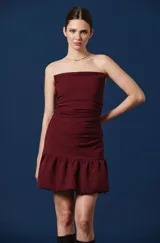 Vestido corto color bordó, con escote palabra de honor, diseño ajustado con frunces laterales y falda con volado en el ruedo.