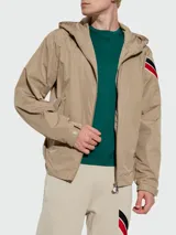 Rompevientos beige con capucha y cierre frontal con cremallera. Tiene bolsillos laterales con cierre de botón a presión y detalle de franjas rojas, blancas y negras en la manga izquierda.