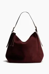 Cartera de hombro de gamuza color bordó, con cierre superior y tirador con detalles de nudos. Cuenta con correa ajustable mediante hebilla metálica en los laterales y diseño estructurado pero flexible.