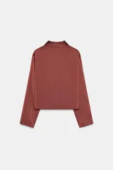Blusa de tejido satinado color bordó, con cuello solapa, manga larga y detalle de abertura drapeada en el pecho. Cuenta con cierre frontal mediante un botón forrado.