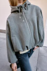 Chaqueta estilo bomber de corte oversize en color marrón oscuro, confeccionada en tejido suave. Presenta capucha con cordones ajustables, cierre frontal con cremallera, mangas largas con puños elásticos y bolsillos delanteros de vivo. El bajo cuenta con detalle de pliegues.