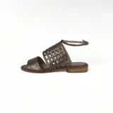 Sandalia plana color peltre, con tiras de cuero entretejidas en el empeine y pulsera con hebilla al tobillo.