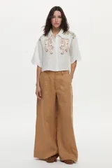 Camisa crop blanca de lino con silueta relajada, cuello clásico y mangas cortas. Presenta bordados de tigres en el frente y detalles ornamentales en los hombros.