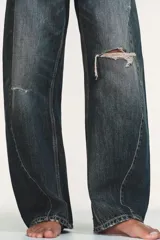 Jeans de tiro medio con diseño baggy balloon, cinco bolsillos y costuras laterales torcidas. Presenta un acabado desgastado con roturas localizadas y cierre frontal con cremallera y botón metálico.