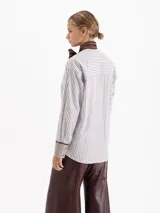 Camisa de poplín con diseño de rayas verticales en tonos azul y blanco. Presenta un cuello prolongado que se anuda en forma de lazo frontal, mangas largas con puños anchos y corte holgado.