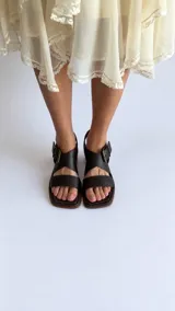 Sandalias de cuero negro con textura croco, punta cuadrada y taco bloque de altura media. Cuenta con tiras cruzadas en el empeine y una hebilla metálica grande decorativa.