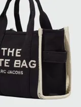 Cartera tipo tote bag de lona color beige, con diseño estructurado y asas superiores. Presenta el logo de la marca estampado en la parte frontal y detalles de cintas laterales con inscripciones.